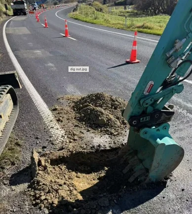 An excavator bucket lifting pavement material from a test pit on a sealed rural highway, with traffic cones separating the work area from the live lane.