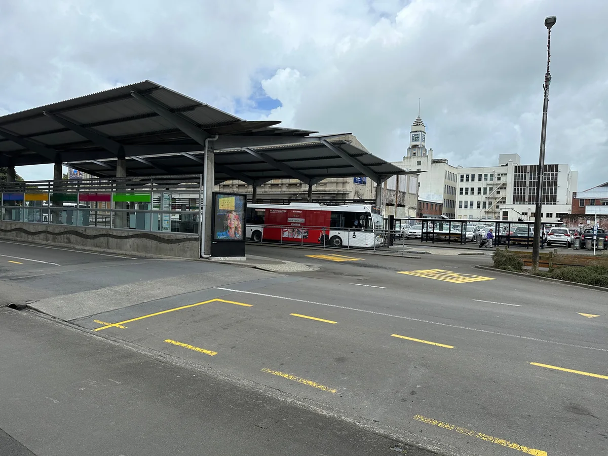 The existing Palmerston North city-centre bus terminal — a covered canopy over bus bays, with a bus at the stand and the city clock tower visible beyond.